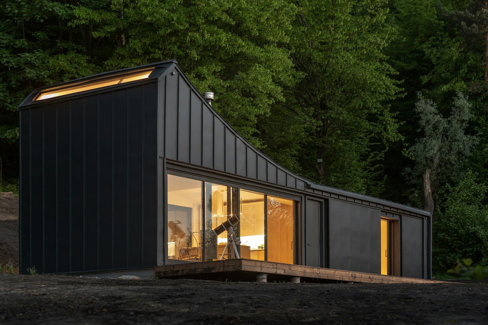 A black timber-clad cabin with a glazed corner lit from inside, among trees