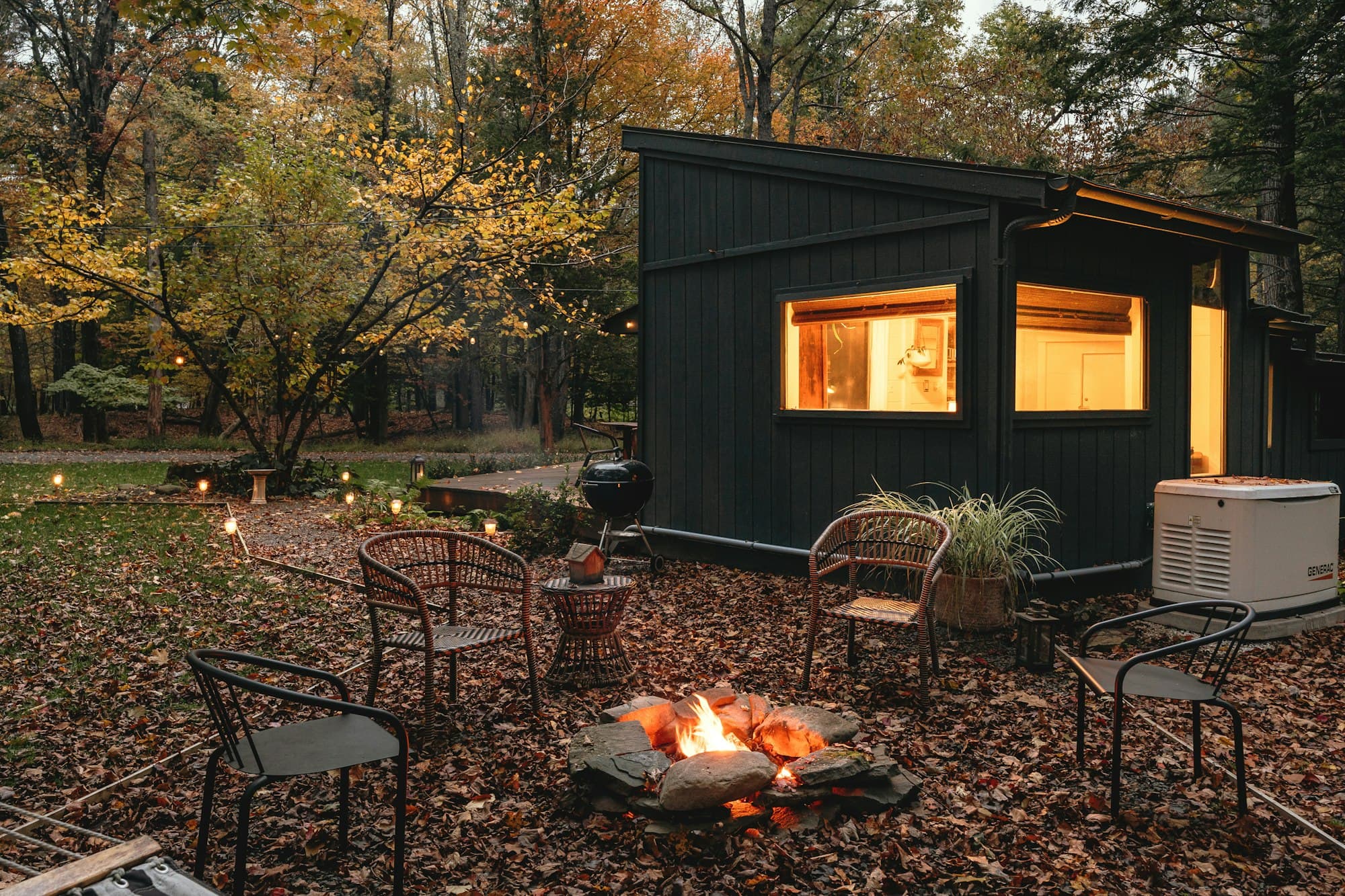A small dark-clad garden room with a lit window, chairs gathered round a fire pit in front