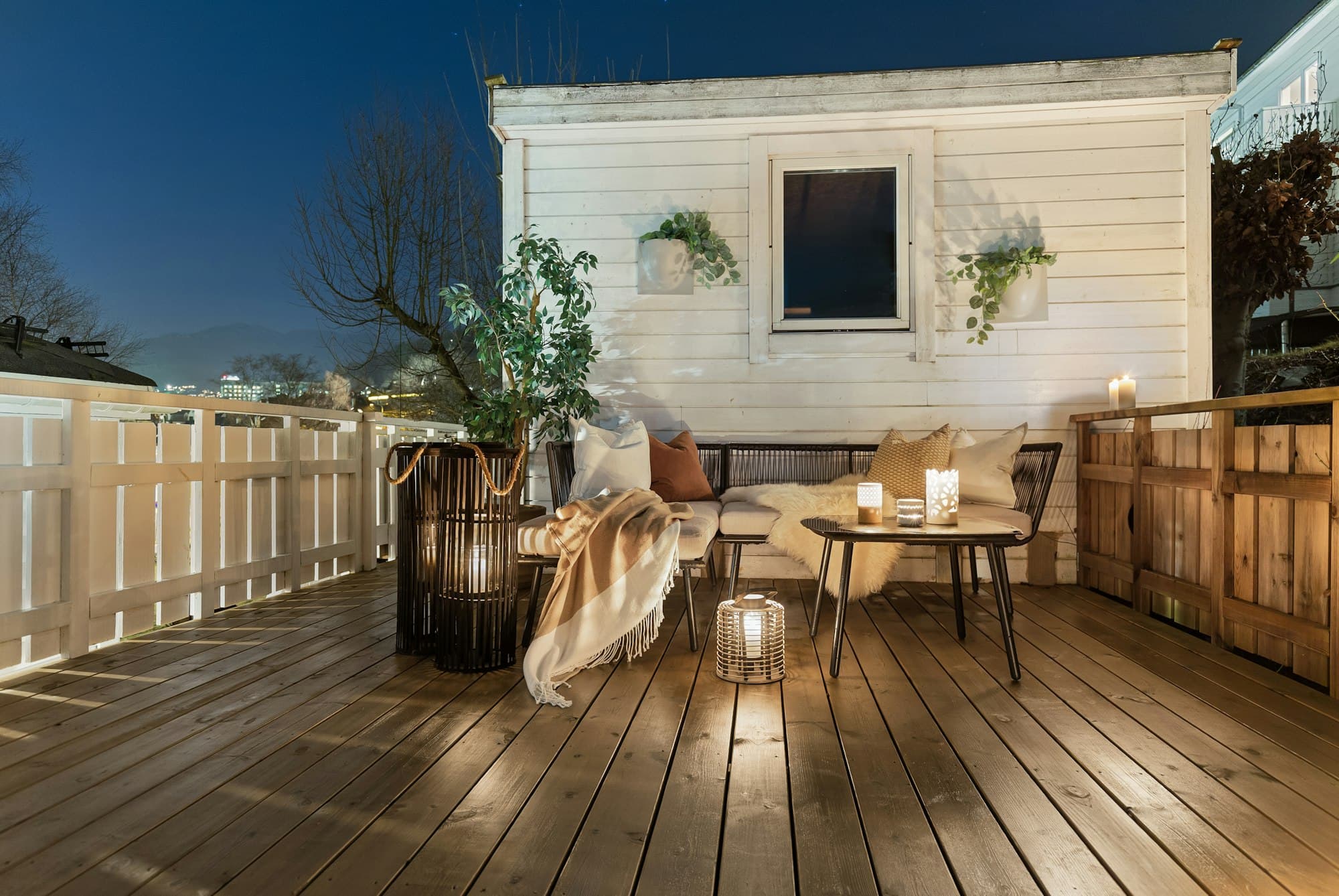 A deck in the evening with a table, lanterns and a blanket outside a white-clad building