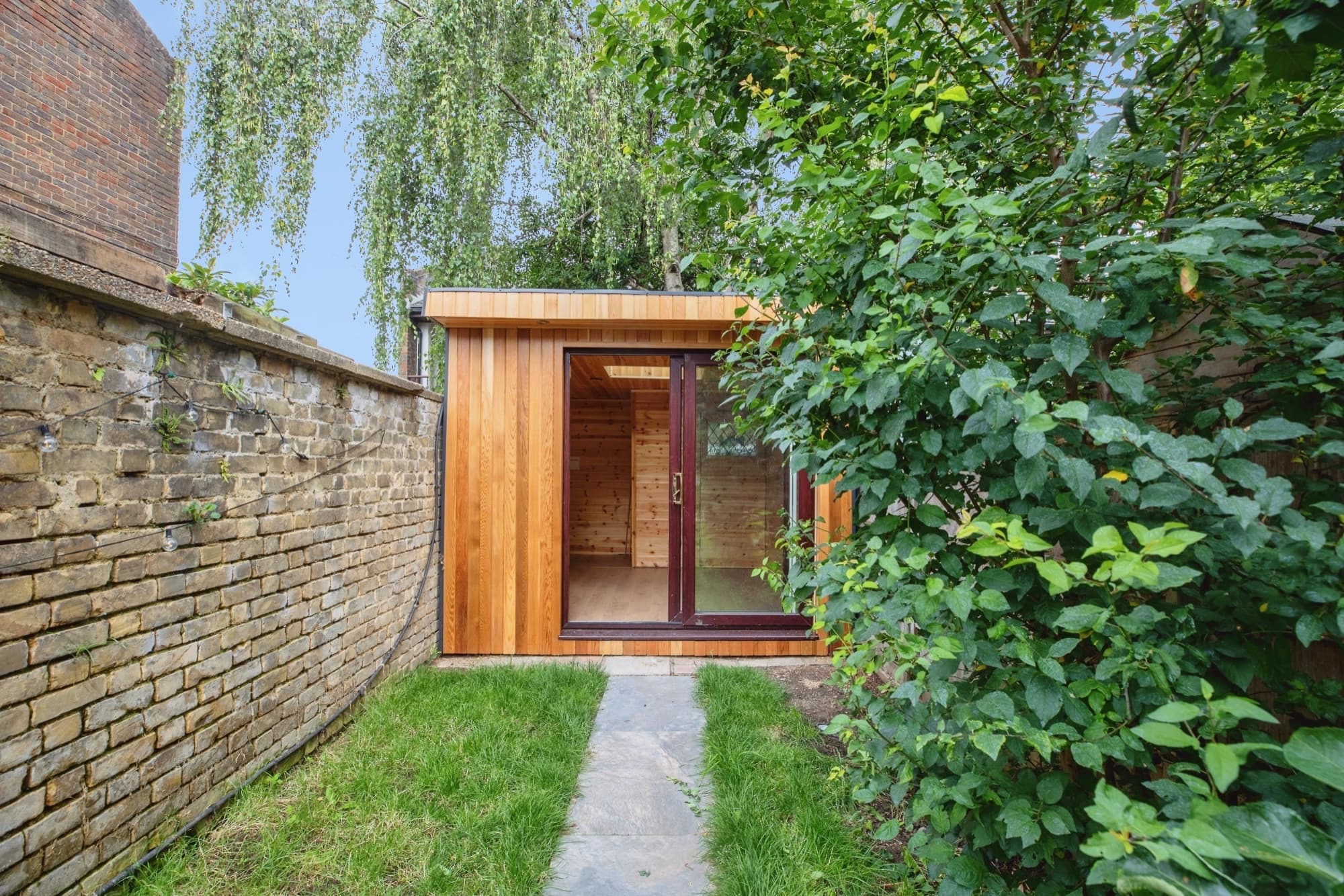 A cedar-clad garden room with glazed sliding doors at the end of a stone path, beside an old brick wall