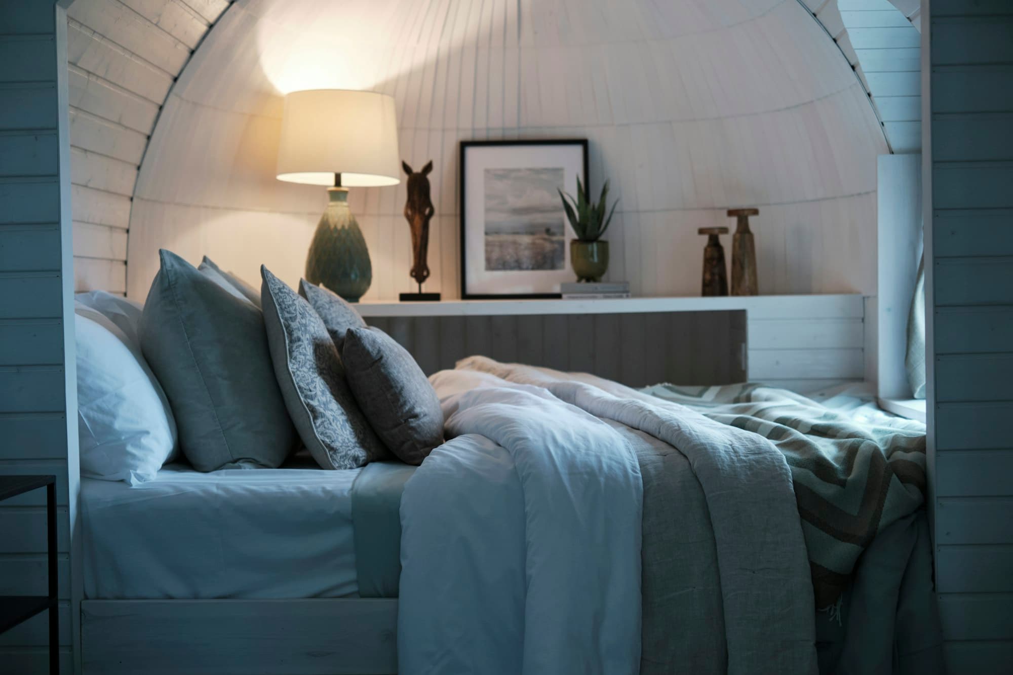 A made bed with blue linen, a bedside lamp and a framed print