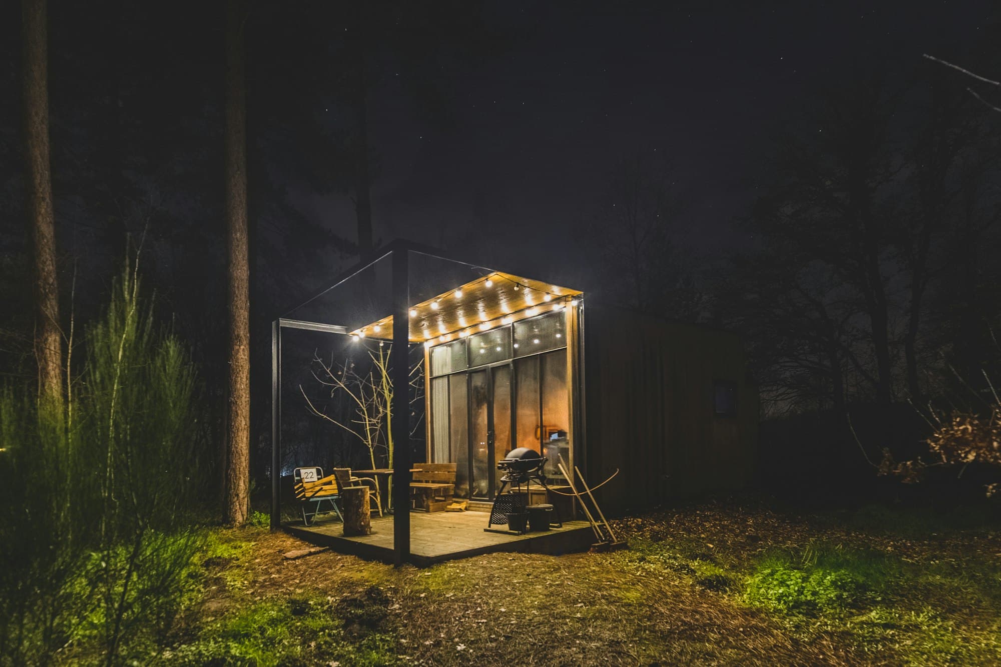 A small glazed garden room lit from inside at night, string lights along the front