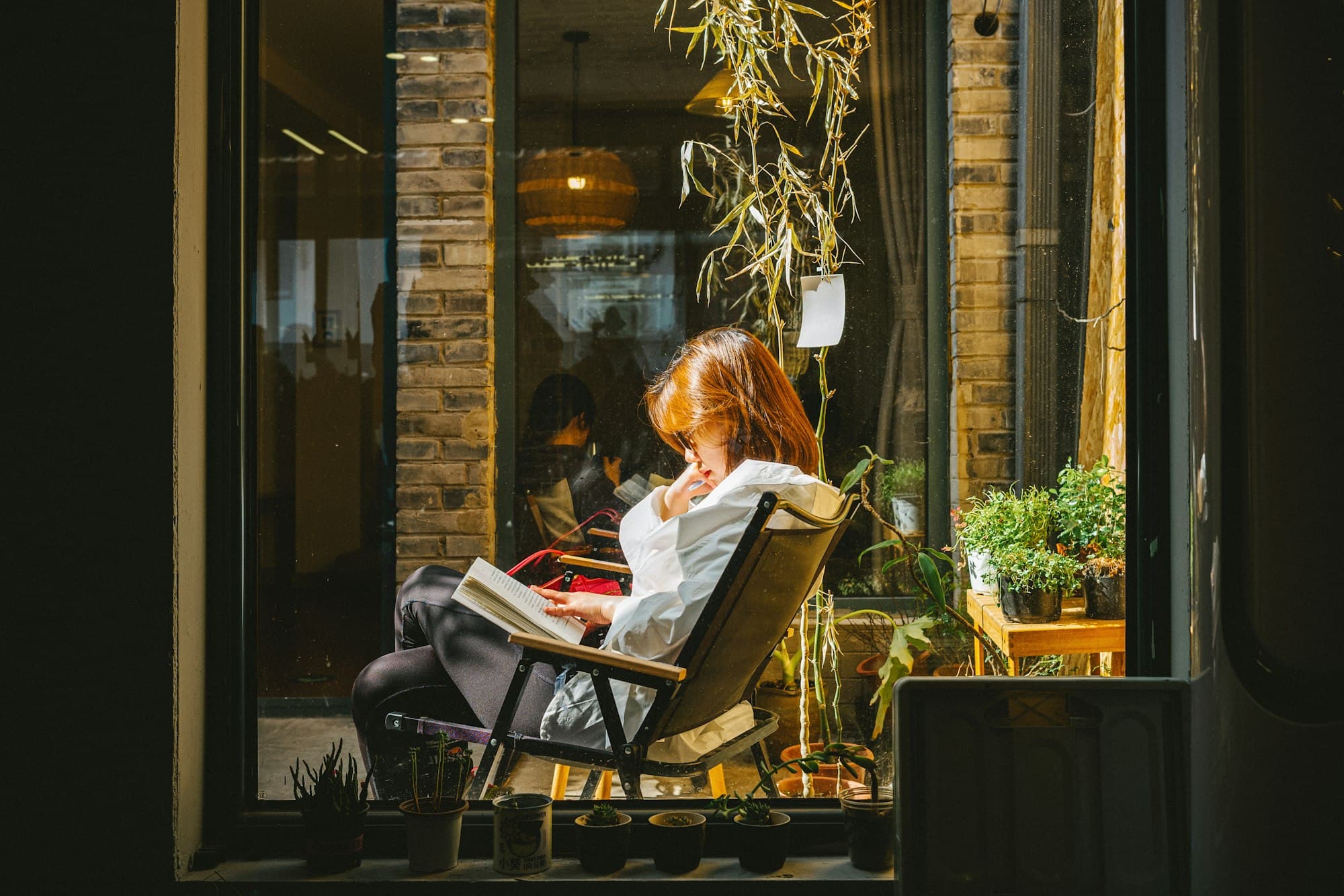 A woman reading in a chair beside glazed doors, garden greenery behind her