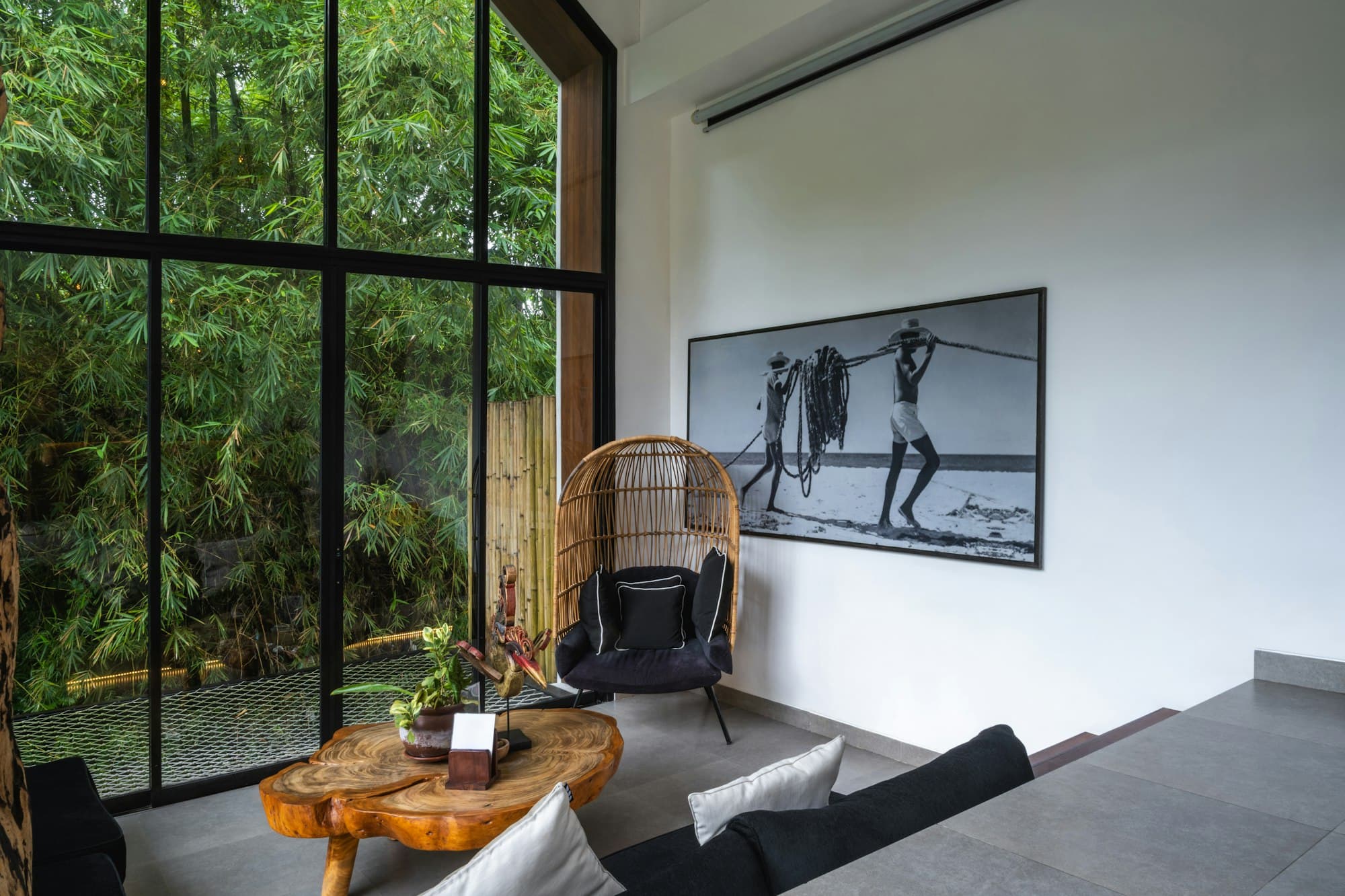 A seating corner with a wicker chair and coffee table beside tall black-framed windows onto trees