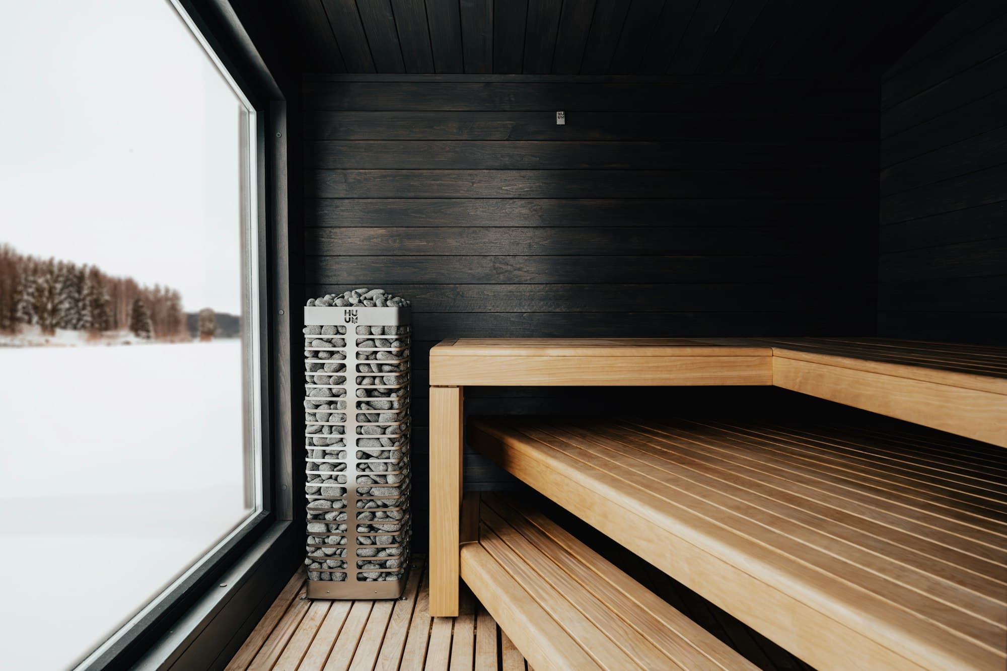 A sauna with a dark wall, a tall heater filled with stones and pale timber benches