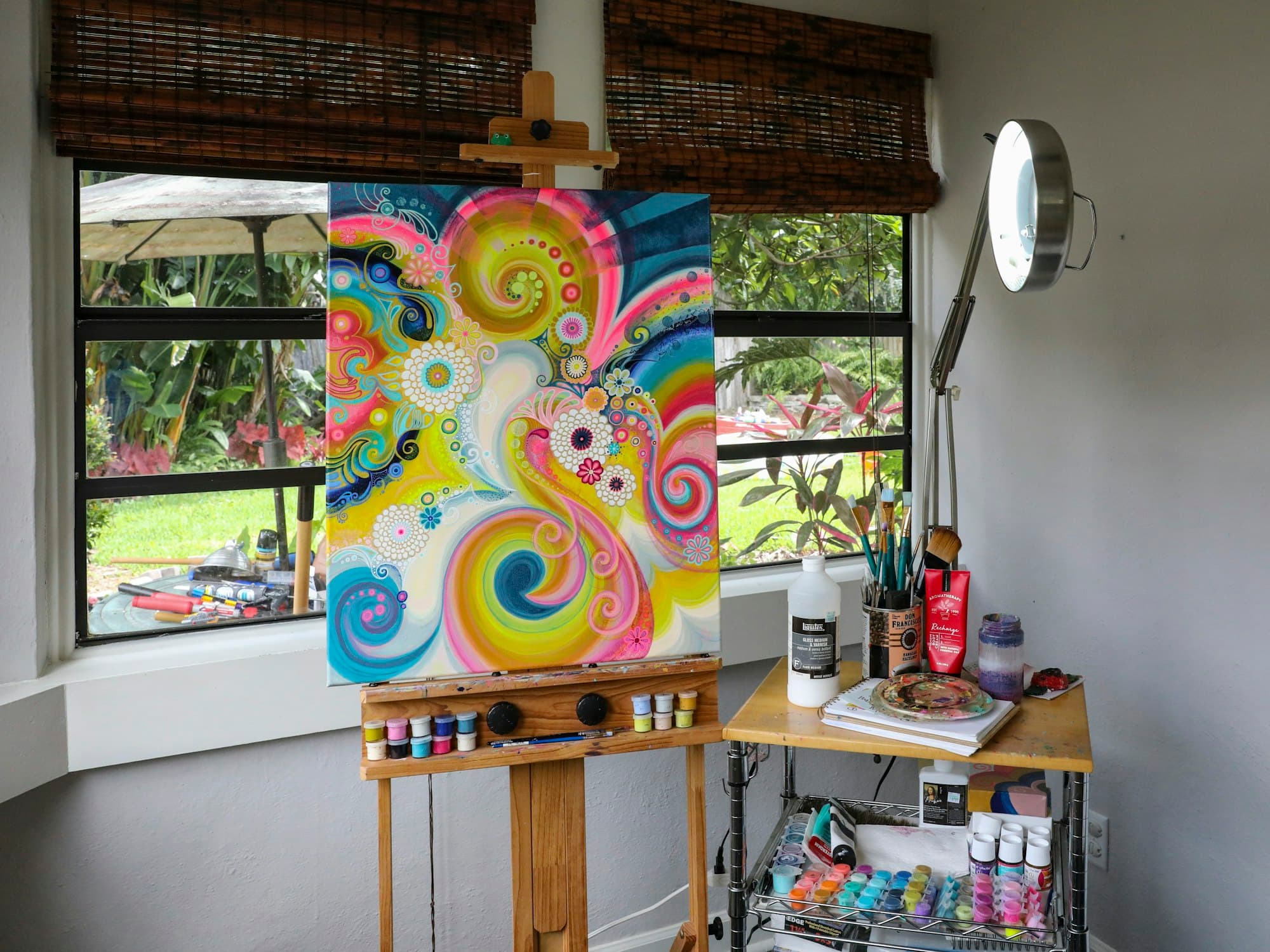 A colourful canvas on an easel beside a window, paints on the table